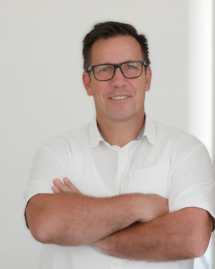 Mann mit Brille und kurzem Haar, der in einem weißen Hemd steht und die Arme vor der Brust verschränkt. Helle, neutrale Umgebung.
