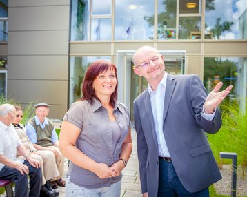 Ein Mann und eine Frau stehen lächelnd im Vordergrund, während im Hintergrund mehrere ältere Männer auf einer Bank sitzen. Die Szene spielt sich vor einem modernen Gebäude mit großen Fenstern und grüner Vegetation ab.